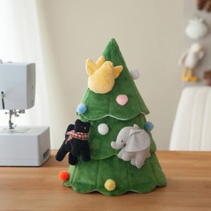 Könnte beinhalten: Ein grüner Weihnachtsbaum aus Plüsch, verziert mit Stofftier-Ornamenten. Der Baum hat einen gelben Vogel, einen schwarzen Bären mit Schal und einen grauen Elefanten. Bunte Pompons und weiße Kugeln sind ebenfalls angebracht.