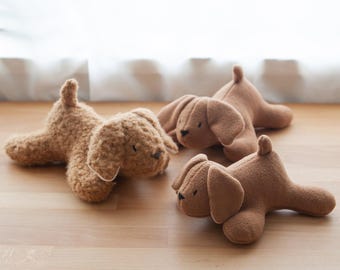 Puede incluir: Cuatro juguetes de peluche con forma de perro en varios tonos de marrón están colocados sobre una superficie de madera. Los perros están tumbados, con orejas caídas y pequeñas colas. Los peluches tienen un aspecto suave y texturizado.