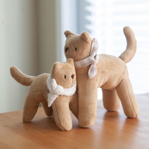 May include: Two tan plush cat toys. One cat is slightly smaller and has a white scarf. The larger cat has a tan and white checked scarf. Both have small, dark brown eyes and are standing on a light brown surface.