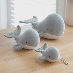 Puede incluir: Tres juguetes de peluche con forma de ballena en diferentes tamaños, cada uno con una parte superior gris suave y una parte inferior blanca. Las dos ballenas más grandes son peluches independientes, mientras que la más pequeña incluye un llavero.