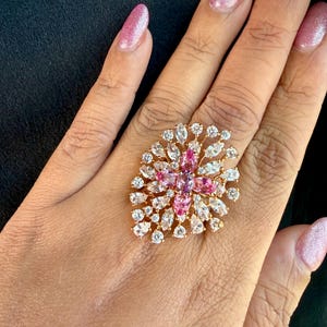 May include: A gold-toned statement ring featuring a cluster of marquise and round-cut clear white stones surrounding a central arrangement of four oval-cut pink stones. The design creates a floral-inspired burst pattern with a metallic gold base.