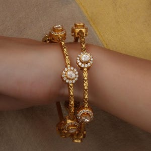 Op de afbeelding: Twee gouden armbanden met ingewikkelde details en sprankelende witte edelstenen. De armbanden hebben een herhalend patroon van diamantachtige stenen in een cirkelvormig ontwerp. Het goud heeft een getextureerd uiterlijk, wat bijdraagt aan de elegantie van de sieraden.