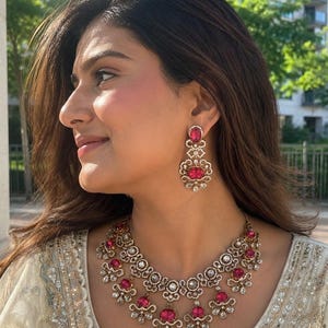May include: A gold-toned necklace and matching drop earring set featuring ornate filigree metalwork, clear pavé-set crystals, and vibrant oval-shaped fuchsia pink stones. The necklace has a multi-tiered design with dangling floral-inspired accents.