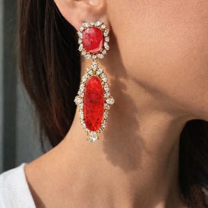 May include: A pair of striking red and gold earrings. Each earring has a square red stone at the top, encircled by small, clear crystals. The lower section features an oval red stone, also framed by clear crystals.