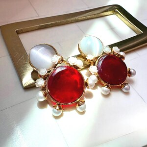 May include: A pair of gold-tone earrings featuring a large circular red stone and a smaller circular white cat's eye stone. The design is accented with small white floral embellishments and tiny white pearls arranged around the red stone base.