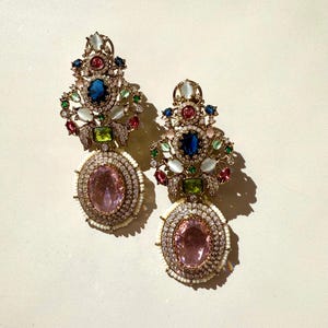 Könnte beinhalten: Ein Paar verzierte Ohrringe mit einem großen ovalen rosa Edelstein, umgeben von kleinen weißen Perlen und klaren Kristallen. Die Ohrringe haben eine goldfarbene Metalleinfassung und sind mit bunten Edelsteinen verziert.