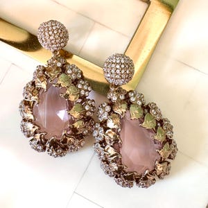 May include: A pair of ornate drop earrings. Each earring features a large, teardrop-shaped, light pink stone surrounded by gold-toned metal leaves and clear crystals. The top of each earring is a small, round, crystal-covered stud.