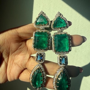 May include: A pair of long drop earrings featuring a heart-shaped green stone at the top, a large rectangular green stone in the center, a small square blue stone, and a pear-shaped green stone at the bottom, all framed by small white stones.