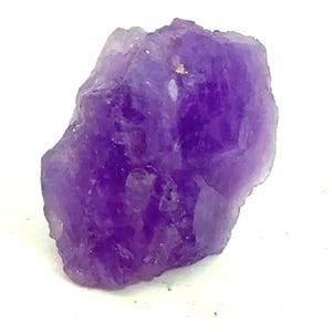 May include: A raw, natural amethyst crystal with a deep purple hue. The crystal has a rough, uneven texture and is set against a white background. This gemstone is a beautiful example of a natural mineral.