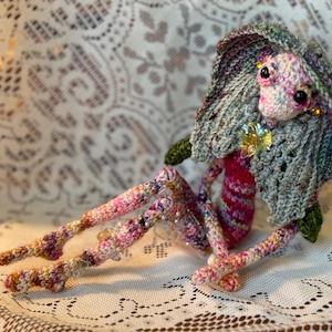 Könnte beinhalten: Eine handgefertigte Häkelpuppe mit einem skurrilen Design. Die Puppe hat ein rosa-violettes Gesicht, große schwarze Augen und einen grauen Schal. Ihre Gliedmaßen sind mehrfarbig, und sie hat einen dekorativen goldenen Akzent. Die Puppe sitzt auf einer weißen Spitzentischdecke.