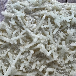May include: A close-up of a light-coloured mineral specimen. The surface is covered with clusters of small, elongated, crystal formations. The crystals are a pale, translucent colour, and the overall texture is rough and uneven.