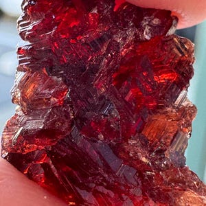 May include: A rough, uncut gemstone in a deep red colour. The crystalline structure is visible, with facets reflecting light. The stone has a rough, natural texture, showing its organic origin. The colour varies from a dark, almost black red to a vibrant, translucent ruby red.