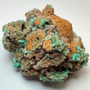 Puede incluir: Espécimen mineral natural con una superficie texturizada. La pieza presenta una mezcla de colores, incluyendo marrón, naranja y verde. Las secciones verdes tienen una apariencia cristalina, contrastando con los tonos terrosos del resto de la roca.
