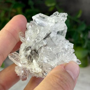 Puede incluir: Un grupo de cristales de cuarzo transparente con terminaciones afiladas y puntiagudas. Los cristales son translúcidos, con algunas áreas que parecen opacas. El cuarzo se sostiene en una mano, con un fondo verde borroso.