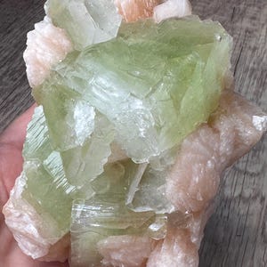 Könnte beinhalten: Ein Mineralexemplar mit großen, durchscheinenden, blassgrünen Kristallstrukturen, die mit undurchsichtigen, hellpfirsichfarbenen Formationen gruppiert sind. Das Cluster weist scharfe, geometrische Kanten und eine abwechslungsreiche, geschichtete Textur auf.