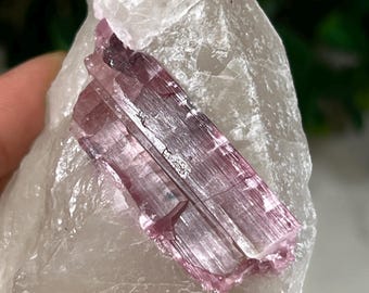 May include: A clear quartz crystal with a pink and white tourmaline crystal embedded inside. The quartz crystal is shaped like a point and the tourmaline crystal is shaped like a rectangle.