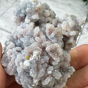 May include: A cluster of small, white, and gray crystals with a slightly pink hue. The crystals are clustered together in a rough, organic shape.