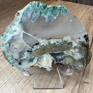 Könnte beinhalten: Eine polierte Achat-Scheibe mit durchscheinenden grauen und weißen Schichten, leuchtend grünen moosartigen Einschlüssen und kristallinen Formationen. Das Mineralexemplar ist auf einem transparenten Acrylständer ausgestellt.