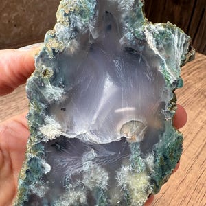 May include: A polished agate slice featuring a translucent, pale lavender-gray center surrounded by a rough, textured border of deep forest green and earthy brown mineral deposits, showcasing intricate crystalline formations and natural inclusions.