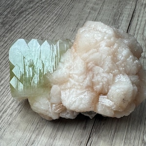 May include: A raw mineral specimen featuring a cluster of pale, translucent, light green crystalline structures on one side, contrasted by a larger, opaque, off-white to light peach-colored crystalline formation with a rough, textured surface.