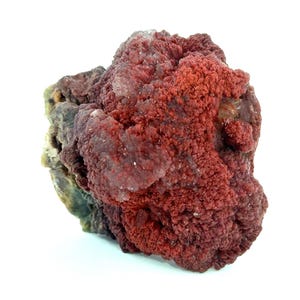 Könnte beinhalten: Nahaufnahme einer Mineralprobe mit rauer, strukturierter Oberfläche. Das Mineral ist überwiegend tiefrot, mit Bereichen von hellerem Rot und einigen durchscheinenden, kristallinen Flecken. Die Basis hat einen grünlich-braunen Farbton.