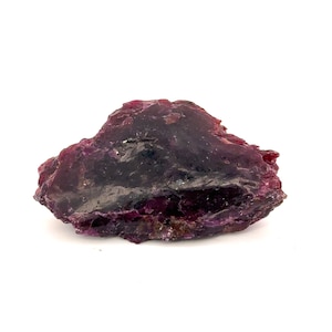 May include: A raw, unpolished gemstone with a deep magenta and dark purple hue. The mineral specimen features an irregular, jagged shape with a translucent, crystalline texture and a glossy, reflective surface across its uneven, natural edges.