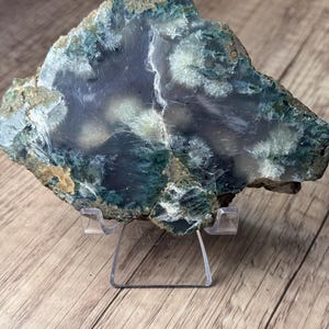 May include: A polished agate slab featuring deep teal and translucent gray tones with white, star-like mineral inclusions throughout the surface. The irregular, rough-edged stone is displayed on a clear, transparent plastic easel stand.