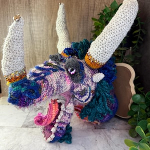 May include: A handcrafted, crochet dragon head sculpture. The dragon features a multicolored design with shades of purple, blue, and pink. It has large, textured horns and a detailed open mouth with a pink tongue. The dragon's eyes are a dark gray with a hint of sparkle.