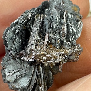 May include: A cluster of gray and silver crystals with a metallic sheen. The crystals are arranged in a fan-like shape and have a rough, textured surface.