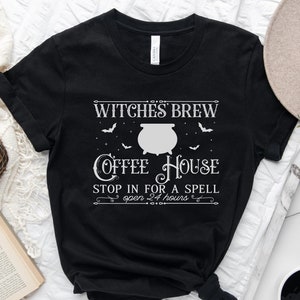 以下が含まれることがあります： 黒いTシャツに白い文字で「Witches Brew Coffee House Stop in for a Spell open 24 hours」と書かれています。テキストは装飾的な枠で囲まれています。デザインには大釜とコウモリがあります。