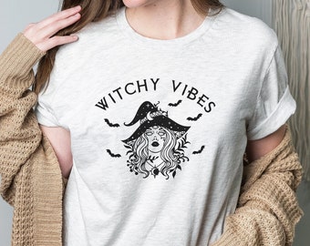 Può includere: Una maglietta bianca con una grafica nera di una strega con un cappello e pipistrelli. Il testo "Witchy Vibes" è sopra la strega.