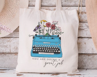 Peut inclure: Sac fourre-tout crème avec une illustration de machine à écrire bleue et des fleurs colorées. Le texte "YOU ARE doing a great job" est imprimé en dessous. Le sac a de longues anses.