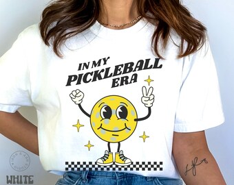 May include: White t-shirt featuring a yellow pickleball character and the text "In My Pickleball Era".