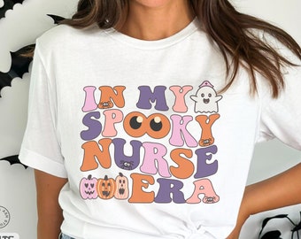 Może przedstawiać: Biała koszulka z napisem "IN MY SPOOKY NURSE ERA" w kolorze różowym, pomarańczowym i fioletowym. Projekt zawiera ducha, dynie i pająki, idealne na Halloween. Koszulka jest w stylu casual i wygodnym.