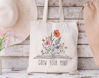 May include: A white canvas tote bag with a floral design and the text "Grow Your Mind". The design features a book with flowers growing out of it.