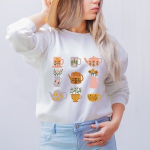 May include: White sweatshirt with a floral pattern of teapots and mugs. The pattern is in shades of pink, yellow, orange, and green.