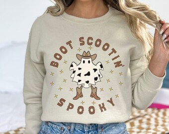 May include: A light beige sweatshirt featuring a graphic of a ghost wearing a cowboy hat and boots. The text "BOOT SCOOTIN' SPOOKY" is printed in brown on the sweatshirt.