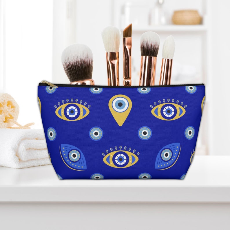 Peut inclure: Une trousse de maquillage bleue &agrave; fermeture &eacute;clair avec un motif r&eacute;p&eacute;t&eacute; d'yeux mal&eacute;fiques dor&eacute;s et bleus. La trousse est pos&eacute;e sur un comptoir blanc avec des pinceaux de maquillage devant elle.