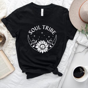 May include: Black t-shirt with a white graphic design of a crescent moon with a sunflower inside and the text "SOUL TRIBE" above it.