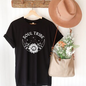 May include: Black t-shirt with a white graphic design of a sunflower inside a crescent moon with stars. The text "SOUL TRIBE" is above the design.