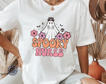 Może przedstawiać: Biała koszulka z różowo-pomarańczowym nadrukiem przedstawiającym ducha z czapką pielęgniarki i napisem "Spooky Nurse".
