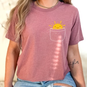 Op de afbeelding: Een mauve t-shirt met een witte zak en een gele lachende zon graphic in de zak.