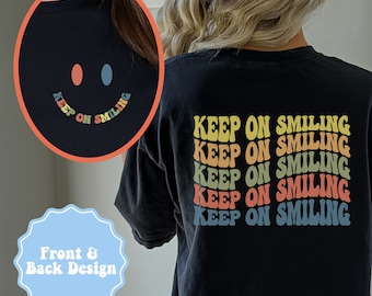 May include: Black t-shirt with a retro smiley face design on the front and the words "KEEP ON SMILING" in a stacked, colorful font on the back. The design includes orange, blue, and yellow hues.