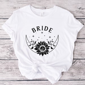 May include: White t-shirt with black text that says "BRIDE" and a black graphic of a crescent moon with a sunflower and stars.