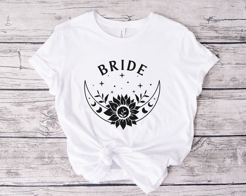 May include: White t-shirt with black text that says "BRIDE" and a black graphic of a crescent moon with a sunflower and stars.
