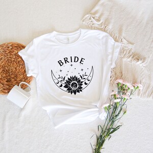 May include: White t-shirt with a black graphic of a crescent moon with a sunflower inside and the word "BRIDE" above it.