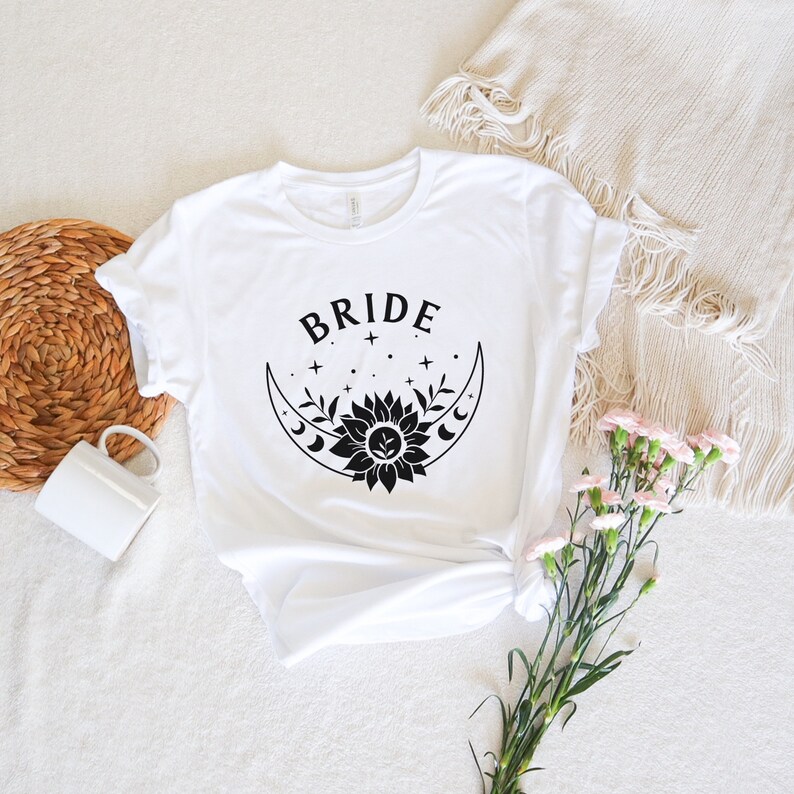 May include: White t-shirt with a black graphic of a crescent moon with a sunflower inside and the word "BRIDE" above it.