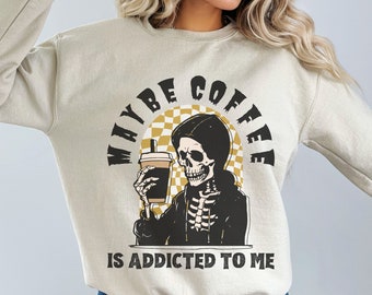May include: A beige sweatshirt with a graphic of a skeleton holding a coffee cup. The graphic is surrounded by a checkered circle with the text "MAYBE COFFEE IS ADDICTED TO ME".