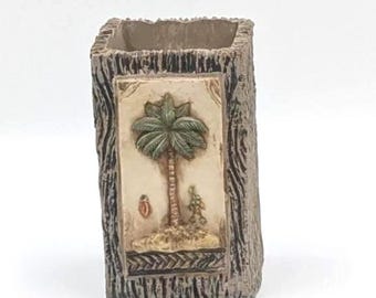 May include: A rectangular pencil holder with a textured brown exterior resembling tree bark. The front features a raised, light-colored rectangular panel depicting a green palm tree on a sandy island, with a small red object and a green figure nearby.
