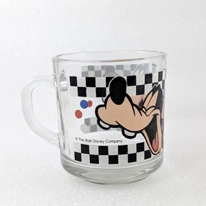May include: A clear glass mug featuring a graphic of the character Goofy with a black and white checkered border. The design includes small red and blue circles and the text The Walt Disney Company printed in black along the lower edge.